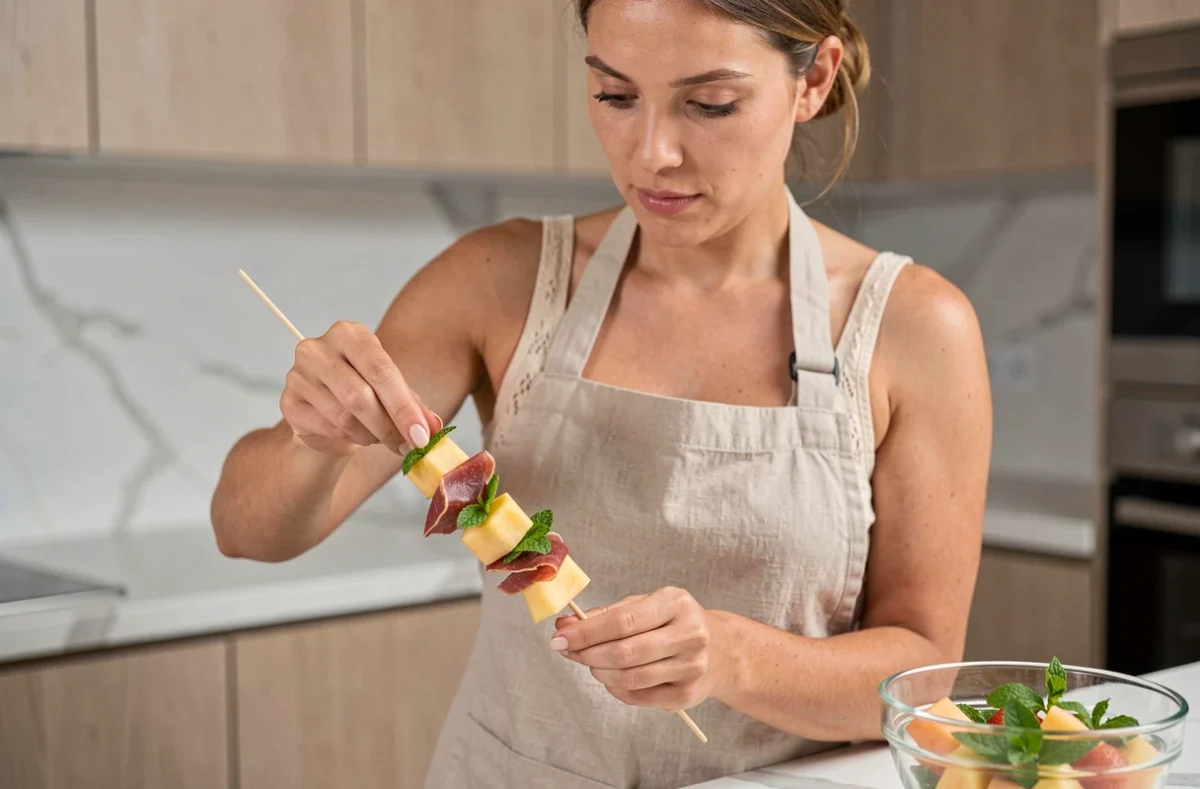Femme préparant des brochettes apéro au melon et jambon cru dans une cuisine moderne la veille d'une fête estivale